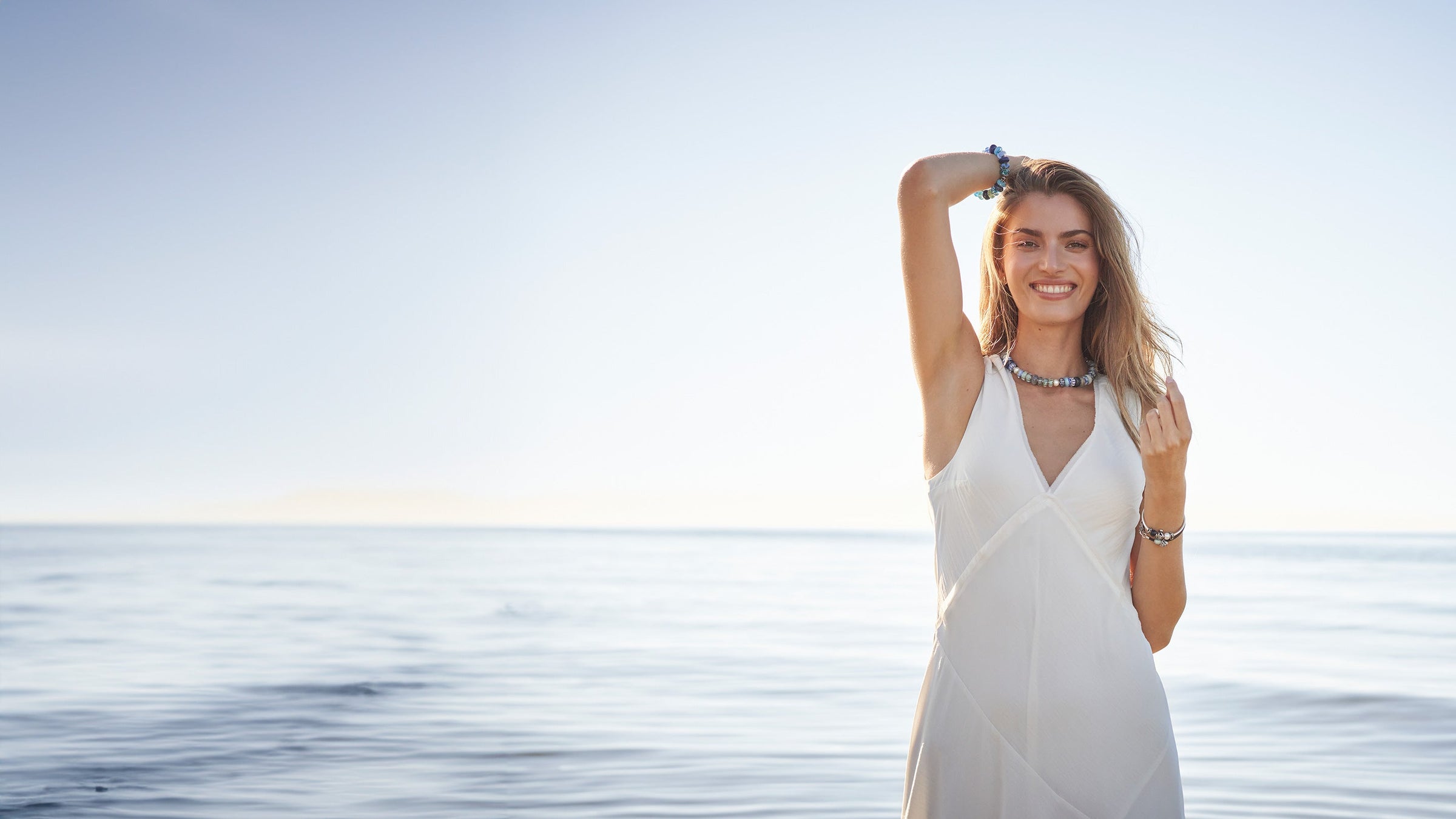 Modèle portant un collier et un bracelet en perles de verre Trollbeads, debout dans l'eau sur une plage, souriant.